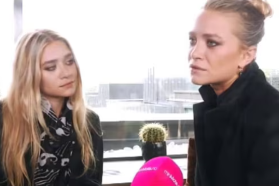 Mary-Kate and Ashley Olsen in a daytime interview with city buildings in the background, one holding a pink microphone as the other listens.