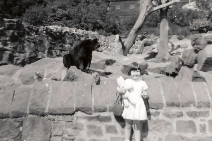 A black-and-white photo of a young girl holding a stuffed animal and smiling in front of a rocky zoo enclosure with a bear standing on a boulder behind her.
