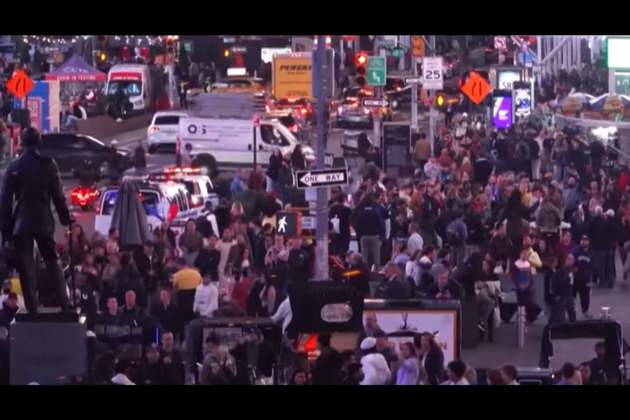 A crowded city street scene filled with people and vehicles, likely in Times Square, New York, with police lights flashing in the background.