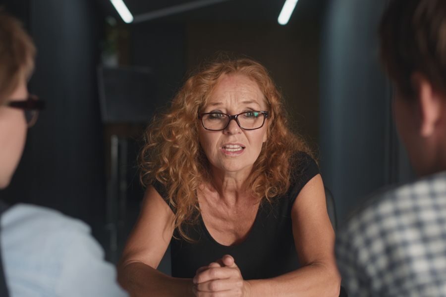 A red-haired woman in glasses leans forward during a serious conversation, speaking to two people seated across from her.