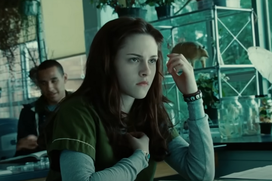 A young woman with long brown hair sits in a classroom, looking serious. She wears a green shirt over a grey top. A classmate is in the background.