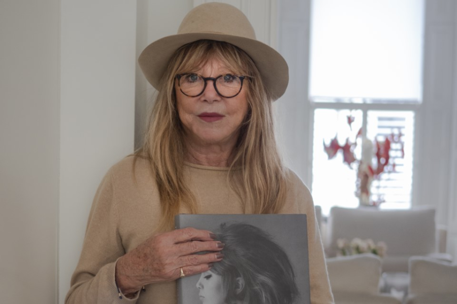 Older Pattie Boyd wearing glasses and a beige hat, holding a large black-and-white portrait of herself from the 1960s.