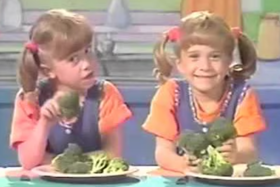 Mary-Kate and Ashley as young children with pigtails, smiling and holding broccoli while seated at a colorful table set.