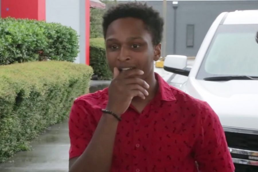 A young man in a red shirt holds his hand near his mouth, reacting with surprise or emotion near a white vehicle in a parking lot.