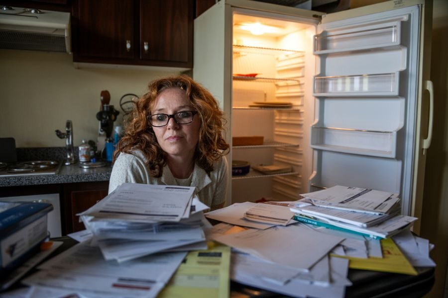 A woman sits at a cluttered kitchen table filled with bills and letters, with an open and nearly empty fridge behind her.