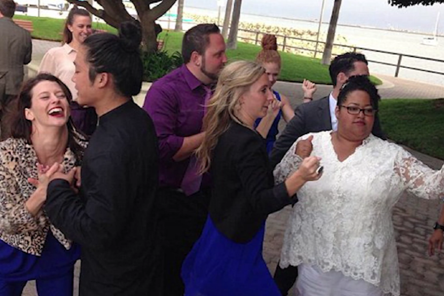 Group of guests, including a bride in white, dancing and smiling at an outdoor wedding with water and palm trees in the background.