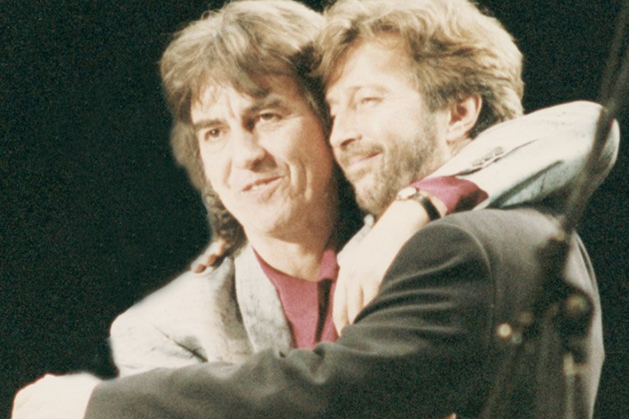 George Harrison and Eric Clapton embrace on stage with warm smiles, both dressed in blazers during a rare public reunion.