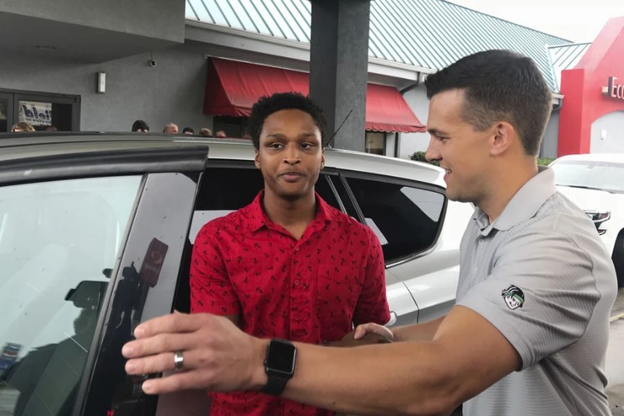 A smiling man in a gray polo shakes hands with a young man in a red shirt next to a car, while others look on in the background.