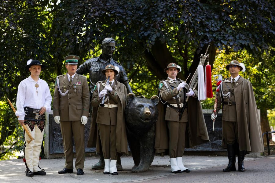 A group of uniformed soldiers and one traditionally dressed man pose solemnly around a bronze statue of Wojtek the bear and a Polish soldier in a park setting.