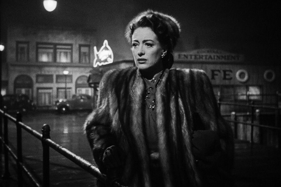 A woman in a fur coat walks down a misty, dimly lit street at night, looking anxious and lost in thought in a classic black-and-white scene.