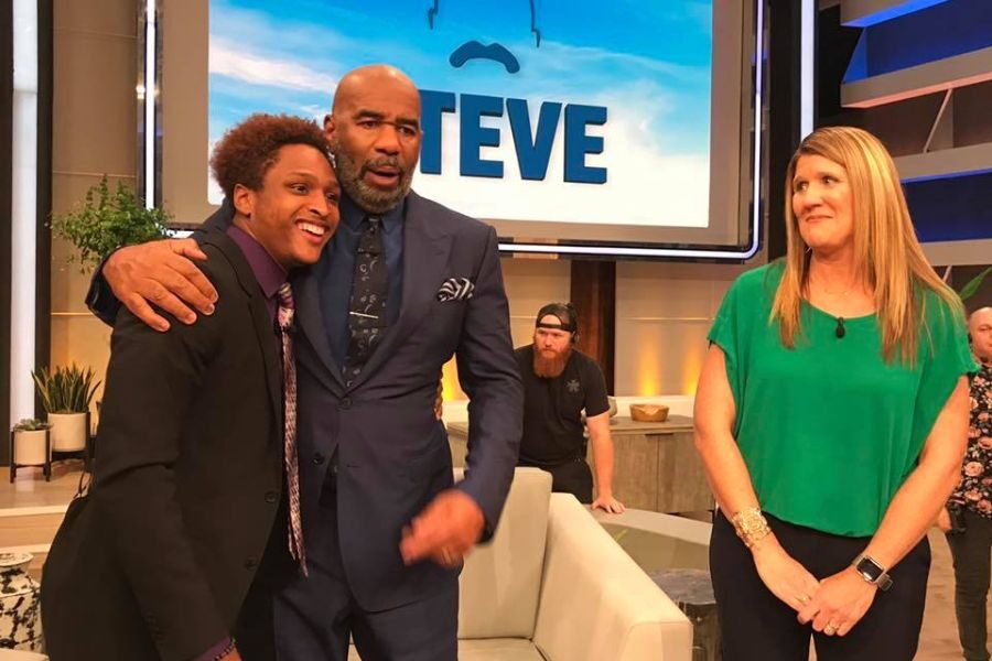 Steve Harvey hugs a smiling young man in a suit while a woman in green stands nearby on the set of the Steve TV show.