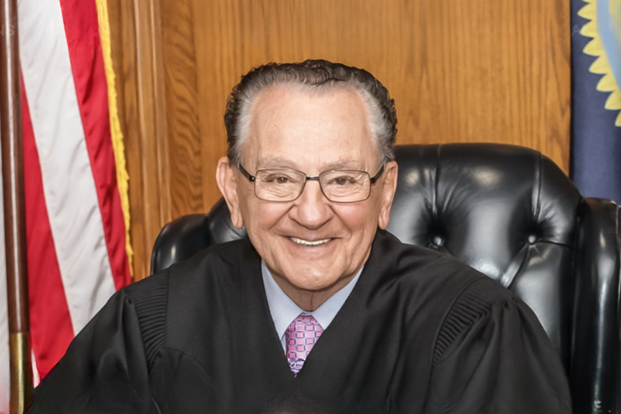 Judge Frank Caprio smiles warmly while seated in his courtroom chair, the U.S. flag and Rhode Island state seal visible behind him.