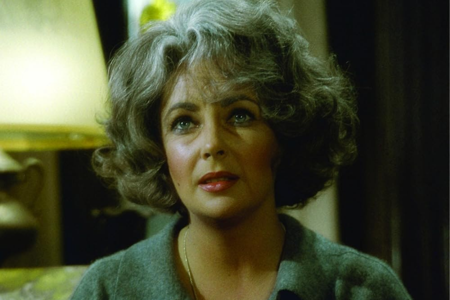A woman with styled gray hair and a soft expression looks slightly upward under warm lighting, wearing a cozy gray top in a vintage interior.