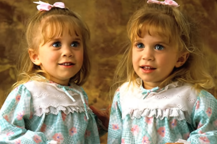 Two young twin girls with blonde hair and blue eyes smile brightly in matching teal gingham dresses with white lace collars and pink bows in their hair. They pose in front of a warm brown studio backdrop.