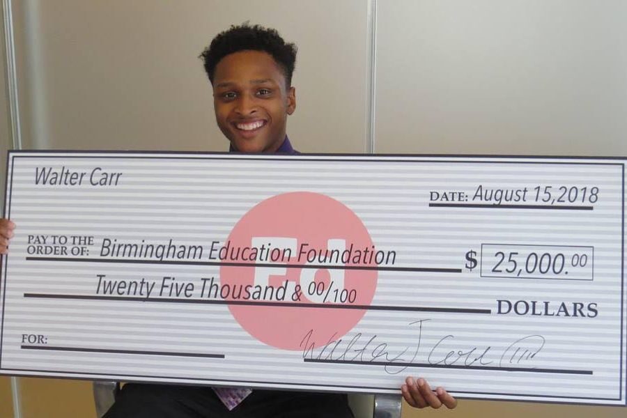 A smiling young man in formal attire holds a large check for $25,000 made out to the Birmingham Education Foundation, dated August 15, 2018.