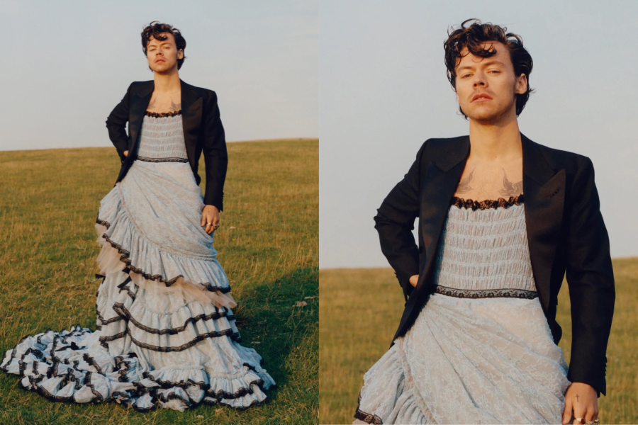 Man standing in an open grassy field wearing a light blue ruffled gown paired with a black tailored blazer. Close-up shows his relaxed stance and natural makeup, blending masculine and feminine style.