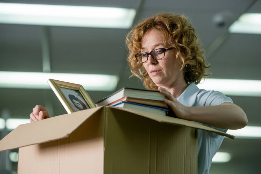 teacher with red hair and glasses solemnly packs framed photos and books into a cardboard box, indicating job loss or resignation.