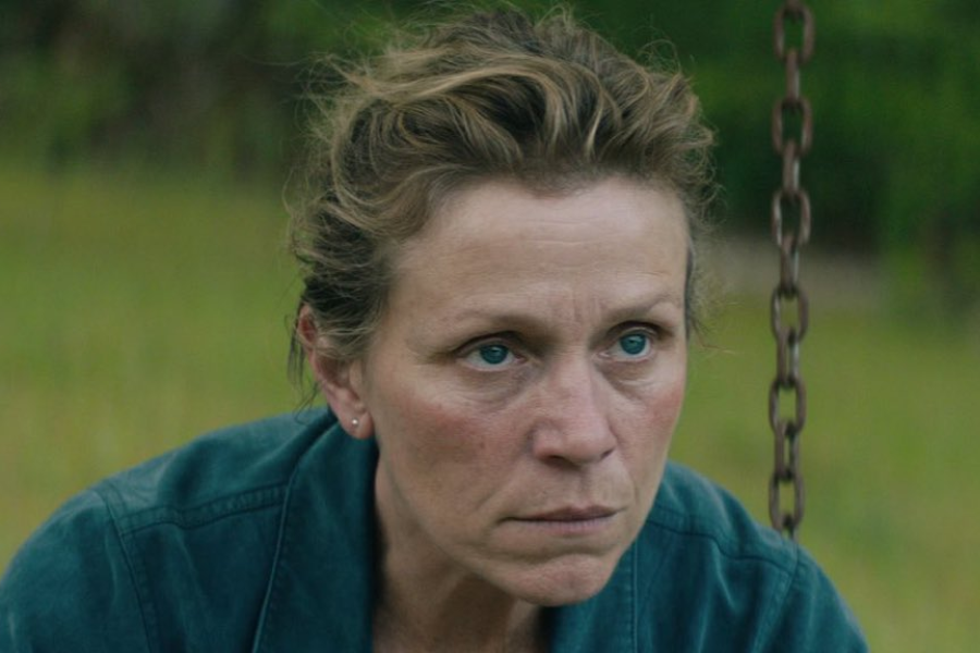 A woman with tied-back hair and an intense stare crouches next to a swing set, wearing a denim jacket in a grassy rural area.