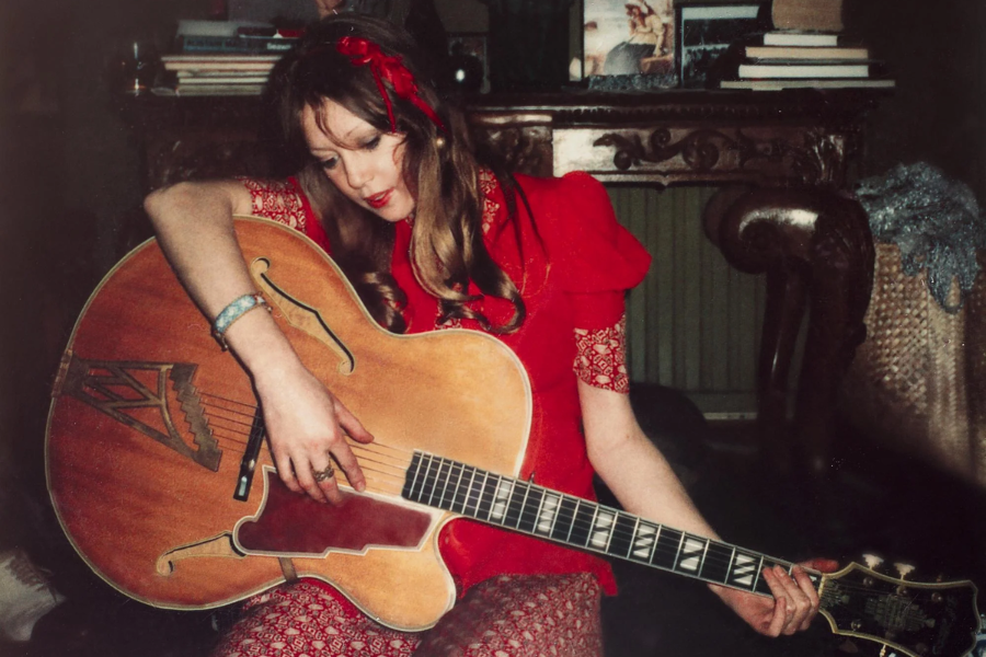 Pattie Boyd in a red outfit gently strums a large vintage guitar in a warmly lit room, looking down with a peaceful expression.