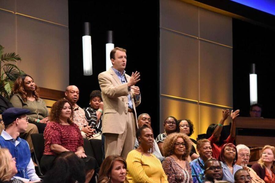 A man in a beige suit stands and speaks among a diverse seated audience during a televised studio recording.