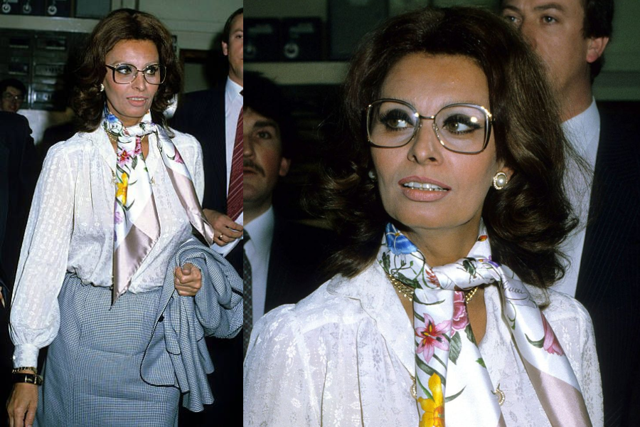 Woman walking through a crowded space in a white blouse with balloon sleeves and a colorful floral scarf, accessorized with oversized eyeglasses and a plaid pencil skirt. Close-up captures her polished hair and dramatic eyewear.