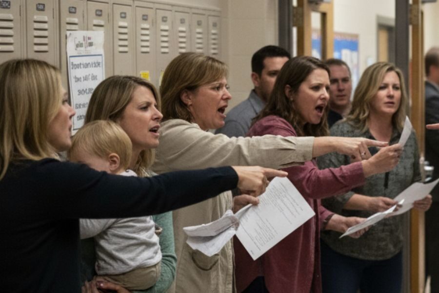 A group of angry parents confront school staff in a hallway, many pointing fingers and holding documents, with lockers in the background.