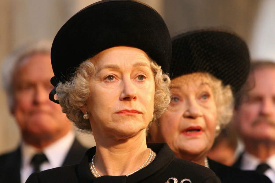 A woman with pearl earrings and short curled gray hair wears a somber black hat and outfit while attending a formal event or ceremony.