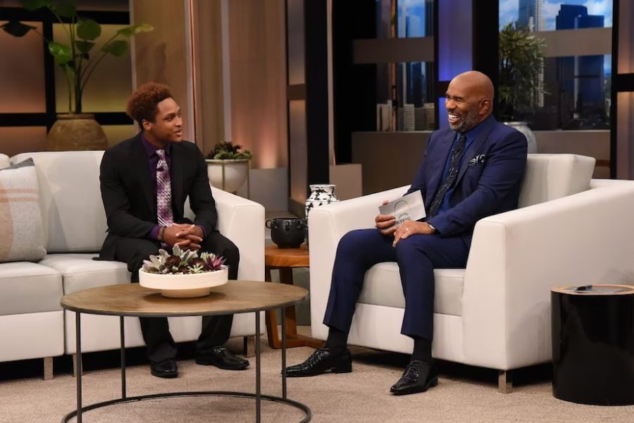 Steve Harvey and a young man in a suit sit on white couches during a televised interview on a modern studio set.