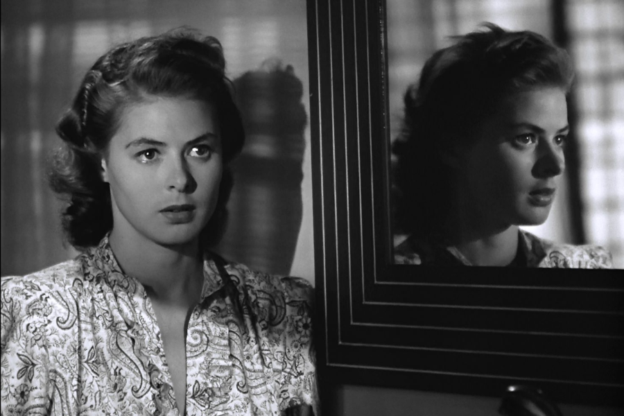 A black-and-white image of a woman staring pensively into the distance, her reflection visible in a mirror beside her.