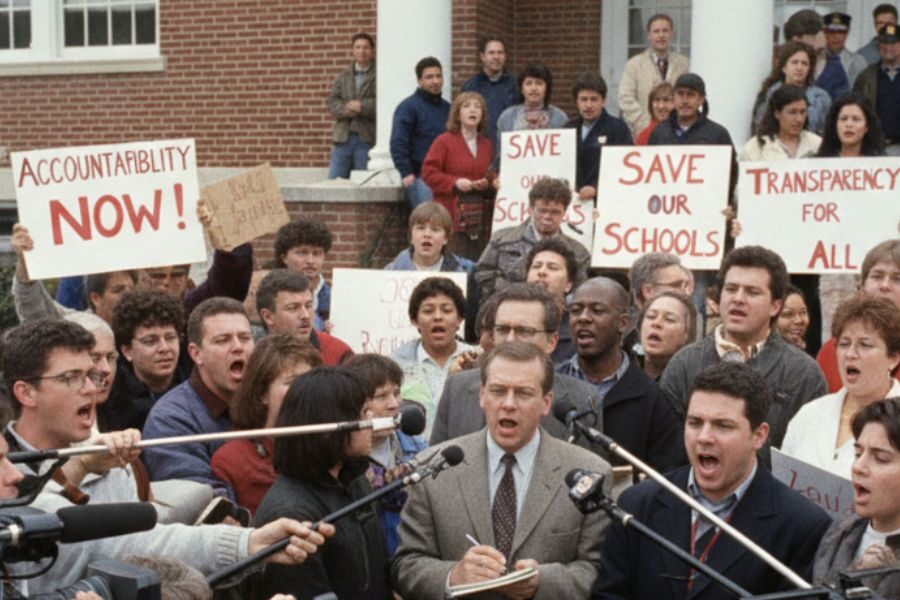 A large crowd of protestors gathers outside a school building holding signs that read “Save Our Schools,” “Accountability Now,” and “Transparency for All.”