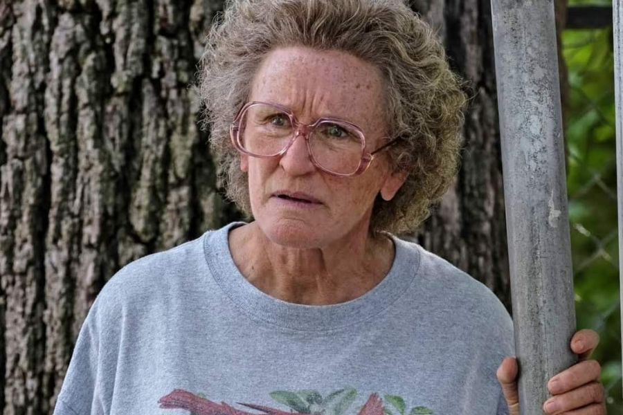 A woman with short, curly gray hair and large pink glasses looks skeptical while gripping a metal pole, wearing a faded t-shirt near a tree.