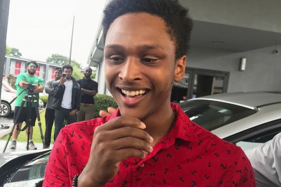 A young man in a red patterned shirt smiles while covering his mouth, as several people in the background record or observe the moment.