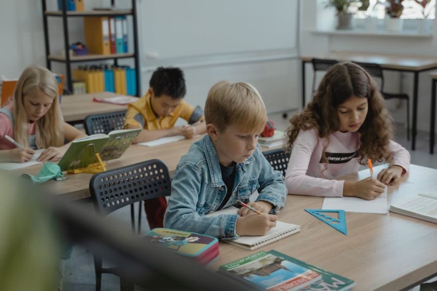 A group of young children focused on writing at their desks in a brightly lit classroom.