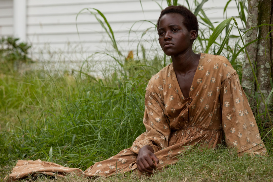 A woman in a faded, brown patterned dress sits on the grass beside a tree, looking off into the distance with a solemn expression. The setting is outdoors, near a white wooden building.