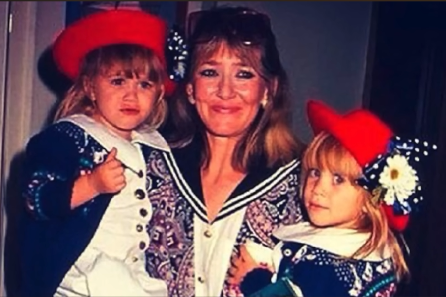 A woman smiles while holding two twin girls in matching sailor-style dresses and red hats with large decorative flowers. One girl looks upset while the other stares quietly at the camera.