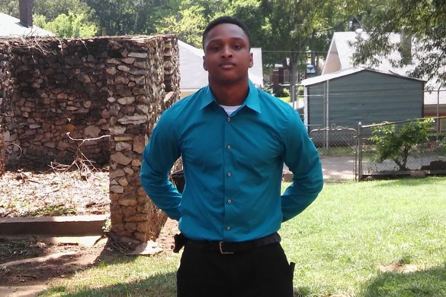 A young man standing confidently with hands behind his back, wearing a teal dress shirt and black pants in a sunny backyard.