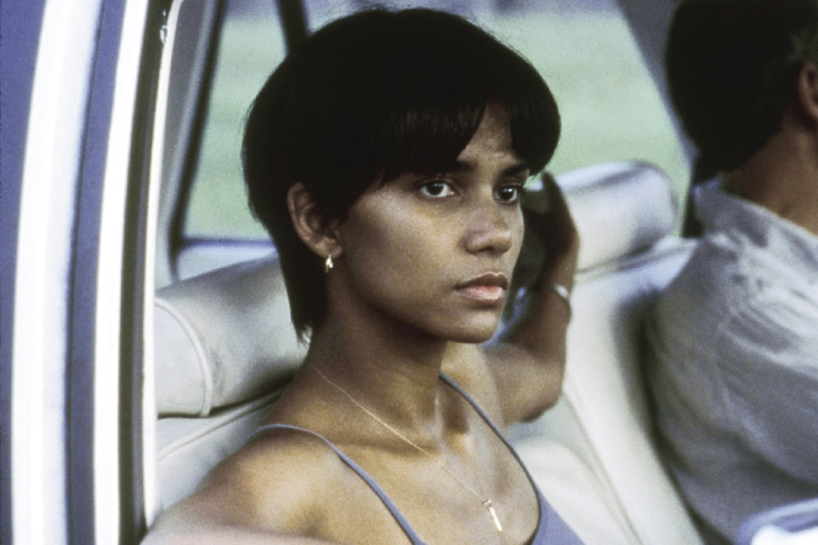 A woman with short dark hair and a serious expression looks out from the passenger seat of a car, wearing a spaghetti strap top and gold jewelry.
