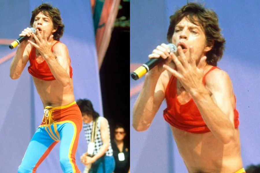 Man mid-performance wearing a red crop top and vibrant color-blocked leggings in red, yellow, and blue while singing into a microphone on stage. Close-up captures his expressive face and animated hand gesture.
