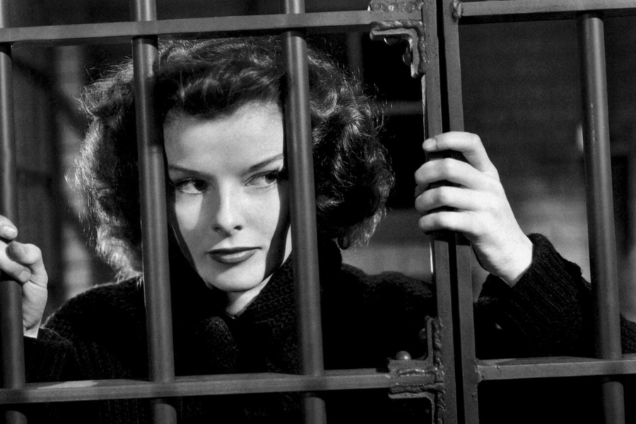 A black-and-white image of a woman with curled hair gripping iron jail bars, giving a sly sideways glance in a dramatic scene.
