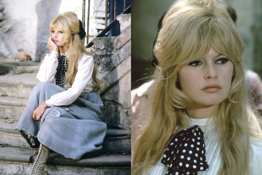 Woman sitting on stone steps in a long grey wool skirt, white blouse, and oversized polka dot necktie with a black bow in her voluminous blonde hair. Close-up highlights her winged eyeliner and classic pout.