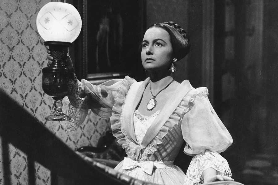 A woman in a voluminous 19th-century gown holds a lit oil lamp as she climbs the stairs with a worried expression in a dark, ornate home.