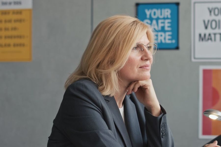 A blonde school administrator in a gray blazer sits thoughtfully with her chin resting on her hand, posters behind her reading “Your Voice Matters” and “Your Safe Here.”