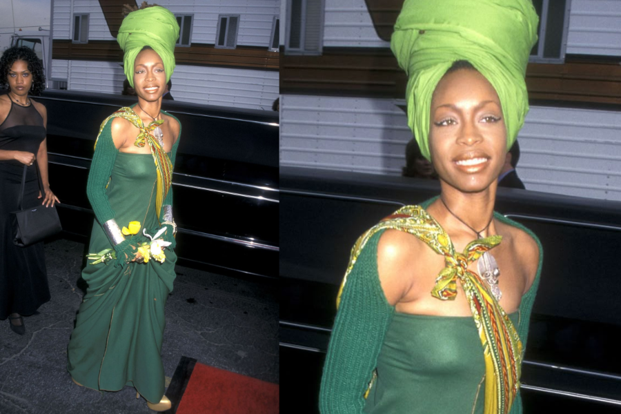 Woman on a red carpet in a forest green gown with yellow accents and a tall green headwrap, holding yellow flowers and wearing metallic gloves. Close-up shows her natural glow and confident smile.
