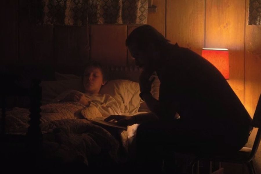 A dimly lit room shows a father sitting beside a child asleep in bed, softly reading a book while a warm lamp glows nearby.