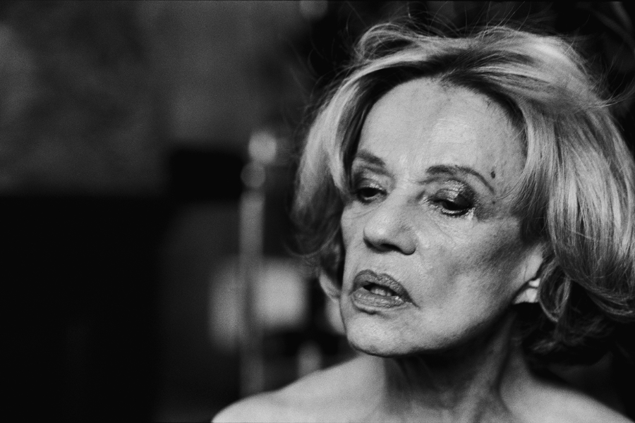 A weary older woman with smudged makeup and tousled hair stares downward, lit dramatically in high-contrast black and white.