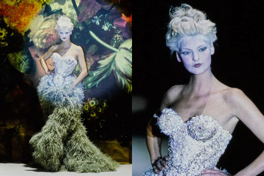 Model on a dimly lit runway in a dramatic mermaid gown with a beaded corset bodice and voluminous feathery skirt, styled with platinum white hair. Close-up captures her icy makeup and sculptural neckline.