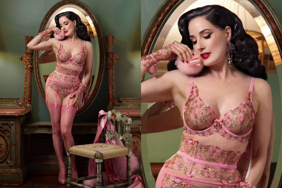 Woman in a pink mesh lingerie set with embroidered flowers and matching gloves, posed in a vintage vanity room with ornate wood furnishings. Close-up highlights her retro curls, red lips, and dramatic earrings.