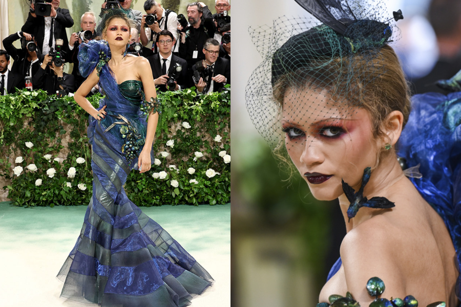 Woman in a one-shoulder iridescent blue and green gown adorned with sculptural florals and dramatic makeup, posing on a flower-lined carpet. Close-up shows her intense eyeshadow, netted fascinator, and floral choker.