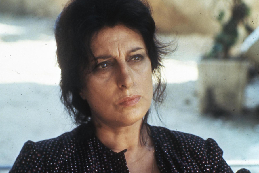 A stern woman with dark hair and furrowed brows stares forward while seated outdoors in the sun, wearing a dotted dress.