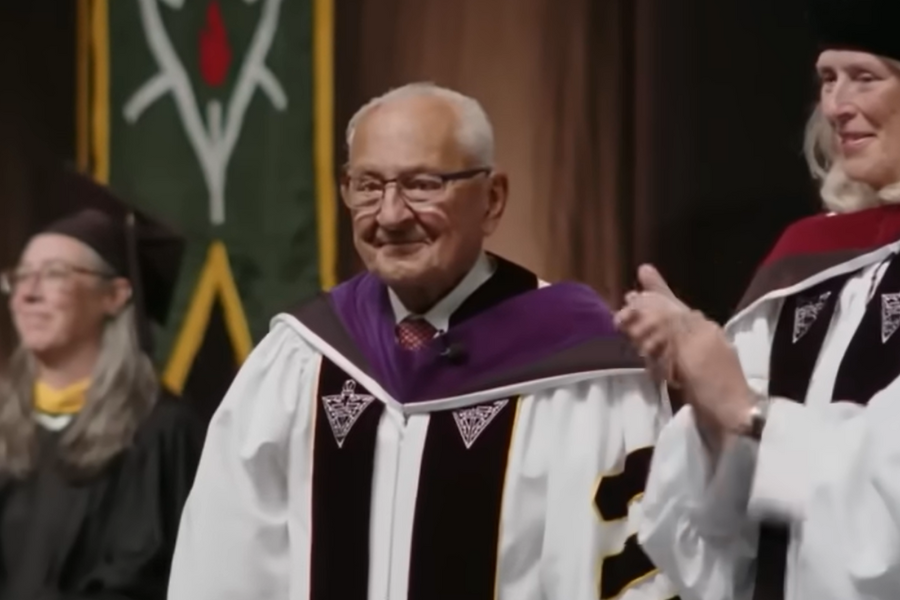 An elderly man in ceremonial academic regalia stands proudly at a graduation ceremony, surrounded by faculty in caps and gowns, receiving an honorary degree.